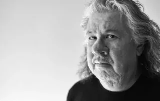 Black and white portrait of Greg Staats, an older man with long hair and a beard, wearing a dark shirt. He gazes to the side with a serious expression, set against a plain background—a compelling moment from the Indigenous Speakers Series.