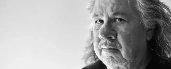 Black and white portrait of Greg Staats, an older man with long hair and a beard, wearing a dark shirt. He gazes to the side with a serious expression, set against a plain background—a compelling moment from the Indigenous Speakers Series.