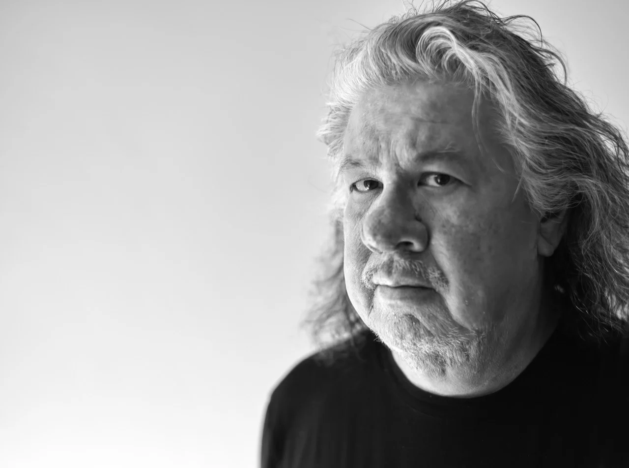 Black and white portrait of Greg Staats, an older man with long hair and a beard, wearing a dark shirt. He gazes to the side with a serious expression, set against a plain background—a compelling moment from the Indigenous Speakers Series.