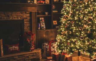 A cozy living room decorated for Christmas, featuring a lit tree adorned with ornaments and lights. Wrapped sustainable gifts sit under the tree. A fireplace is on the left, with shelves displaying various decorative items.