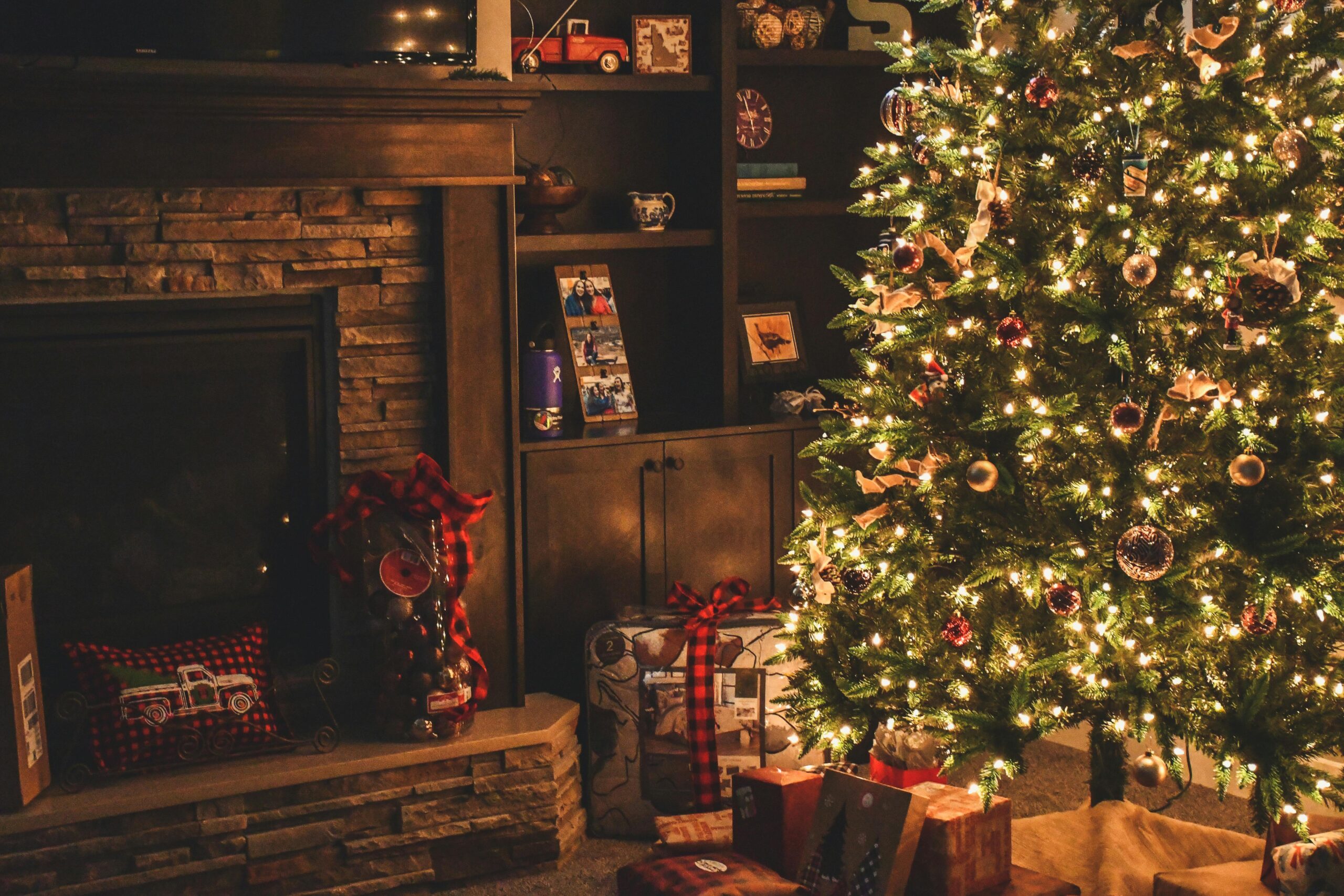 A cozy living room decorated for Christmas, featuring a lit tree adorned with ornaments and lights. Wrapped sustainable gifts sit under the tree. A fireplace is on the left, with shelves displaying various decorative items.