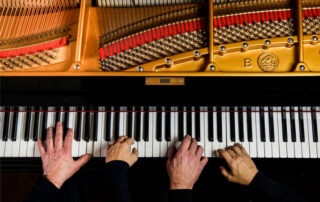 Top view of four hands performing a piano duet on one piano. Two hands grace the left side while another pair dances on the right, set against a backdrop of opened strings and hammers, revealing an intricate tapestry of colorful components within.