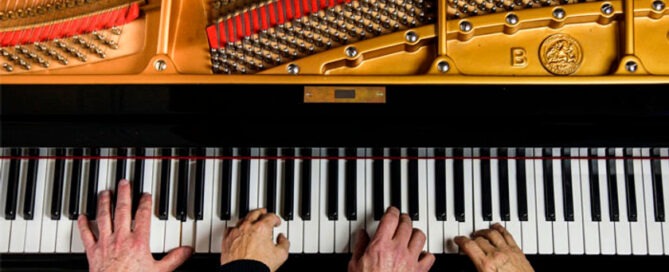 Top view of four hands performing a piano duet on one piano. Two hands grace the left side while another pair dances on the right, set against a backdrop of opened strings and hammers, revealing an intricate tapestry of colorful components within.