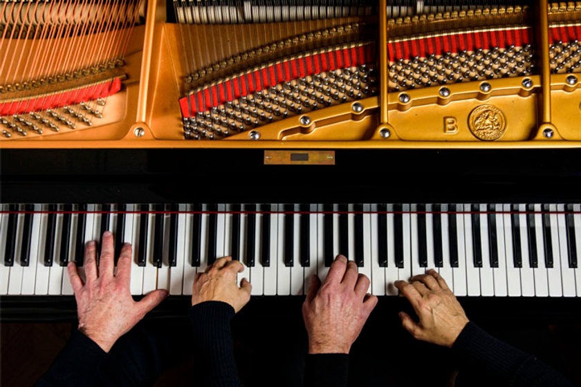 Top view of four hands performing a piano duet on one piano. Two hands grace the left side while another pair dances on the right, set against a backdrop of opened strings and hammers, revealing an intricate tapestry of colorful components within.