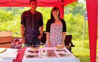 A man and a woman stand smiling under a red canopy at an outdoor event, representing the spirit of the Indigenous community. They are behind a table displaying brochures and items, with lush greenery visible in the background.