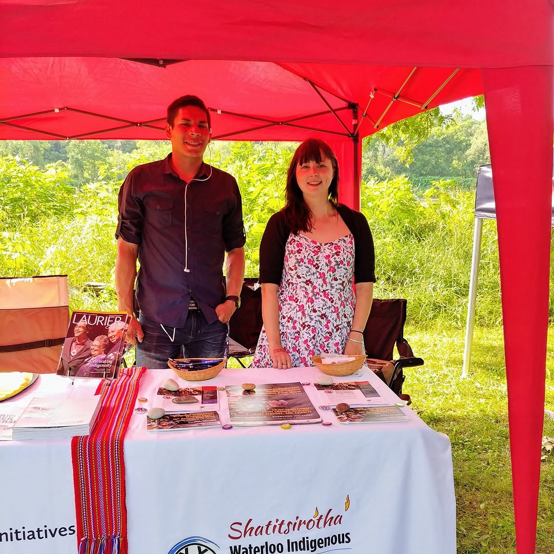 A man and a woman stand smiling under a red canopy at an outdoor event, representing the spirit of the Indigenous community. They are behind a table displaying brochures and items, with lush greenery visible in the background.