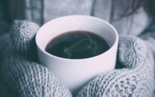 A person wearing gray knitted gloves holds a steaming white cup of coffee, offering solace from the winter blues. The focus is on the cup, with a cozy sweater visible in the background, suggesting a warm and comforting atmosphere perfect for staying well during chilly days.