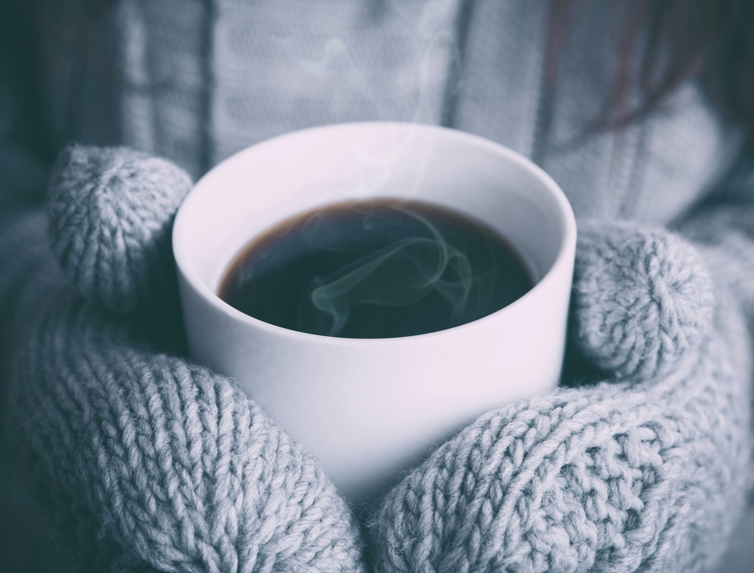 A person wearing gray knitted gloves holds a steaming white cup of coffee, offering solace from the winter blues. The focus is on the cup, with a cozy sweater visible in the background, suggesting a warm and comforting atmosphere perfect for staying well during chilly days.