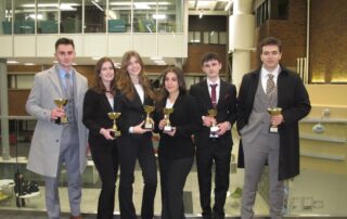 Six people in business attire stand indoors, holding trophies and smiling. They are posing together in front of a large window of the historic urban interior with high ceilings, celebrating their success at the invitational event.