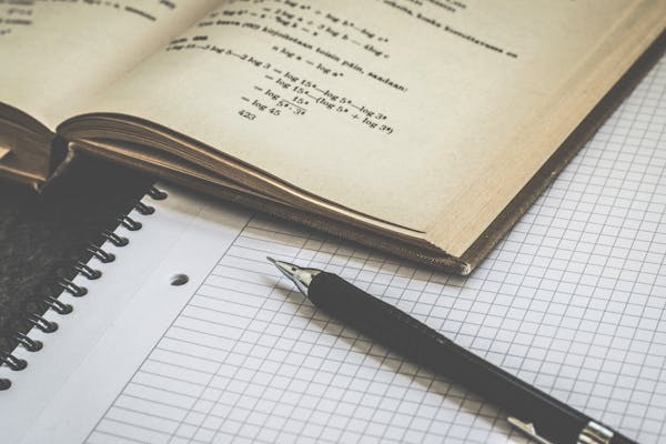 An open book with mathematical equations is beside a pen resting on a stack of graph and lined paper notebooks. The scene suggests exam preparation, with intense study or work involving calculations and note-taking, perfect for tackling the MCAT.