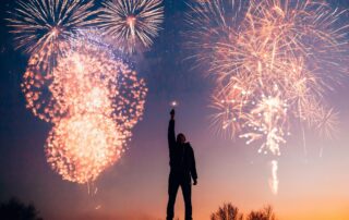 Silhouette of a person with an arm raised, standing against a backdrop of colorful fireworks illuminating the evening sky, celebrating achievable New Year's resolutions. The horizon glows with the fading light of sunset, and trees are visible in the distance as we step towards 2025.