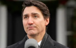 Under the cool sky, a person with short dark hair and clad in a dark coat speaks passionately into the microphone outdoors, as onlookers ponder Trudeau’s resignation.