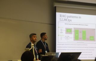 Two presenters stand beside a projected slide titled "RAG patterns in LLMOps" at Geese Hacks, a student-run hackathon. The slide includes diagrams and text about data processing. The room has a whiteboard and various audio-visual equipment. One presenter gestures energetically while speaking.