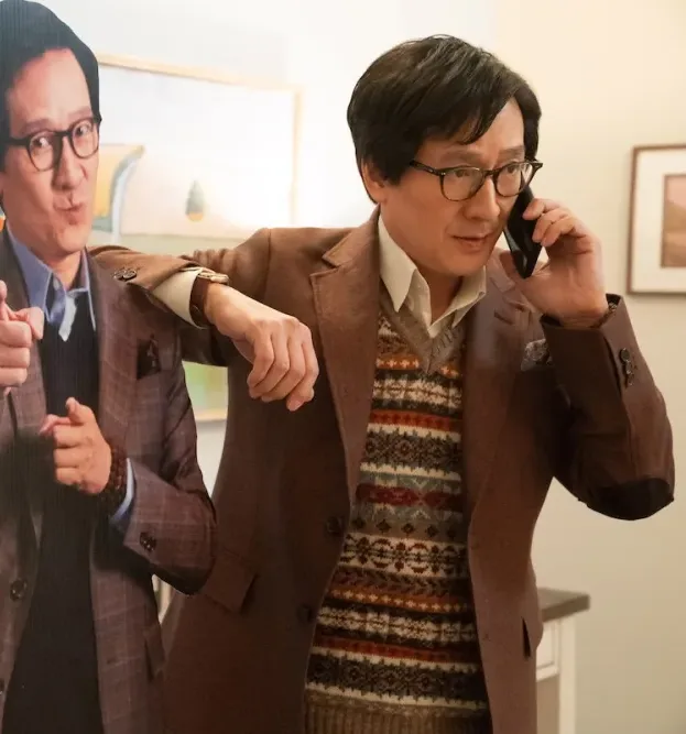 A man in a brown suit and glasses talks on his phone, standing beside a cardboard cutout of himself pointing. The display reads "Frontier Realty" with "Want Home for You!" in bold text. It's reminiscent of a quirky Valentines flick scene, all set in a warmly lit room.