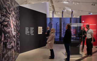 Visitors in a museum hall observe a large artwork depicting intertwined figures. The ROM exhibit features text panels and informational displays, while modern lighting illuminates the room with more exhibits in the background.