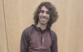 A person with curly hair, resembling Damian Mikhail, smiles while standing against a light wooden background. They are wearing a maroon long-sleeve shirt and a necklace, evoking the poised demeanor of someone ready for the WUSA presidency in 2025-2026.