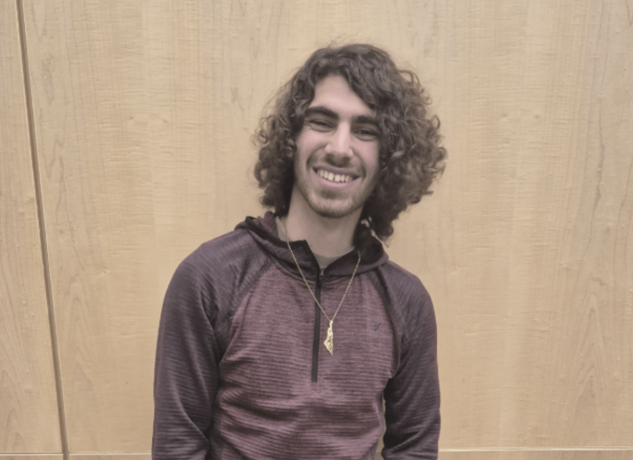 A person with curly hair, resembling Damian Mikhail, smiles while standing against a light wooden background. They are wearing a maroon long-sleeve shirt and a necklace, evoking the poised demeanor of someone ready for the WUSA presidency in 2025-2026.