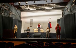 A theater stage set with a minimalist design features five people spaced out, wearing everyday clothing. A couch is placed centrally on an elevated platform. The surrounding area is dark, and unoccupied theater seats are visible in the foreground.