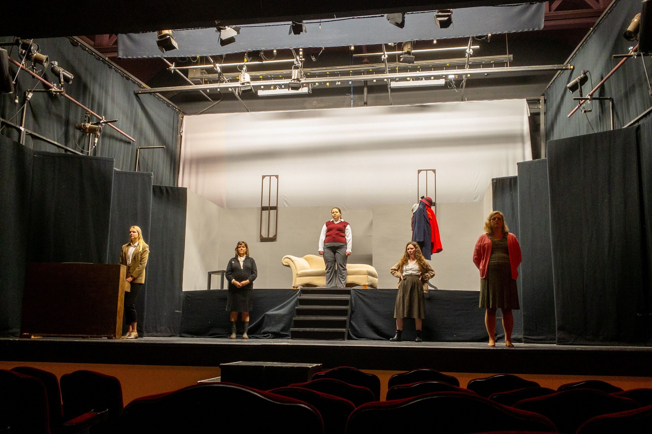 A theater stage set with a minimalist design features five people spaced out, wearing everyday clothing. A couch is placed centrally on an elevated platform. The surrounding area is dark, and unoccupied theater seats are visible in the foreground.