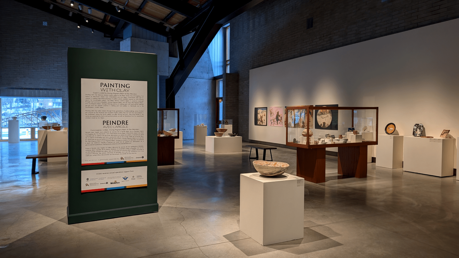 Glass & Clay Gallery presents the "Painting with Clay" exhibit, featuring diverse pottery pieces on pedestals and in glass cases. The walls, adorned with framed Indigenous art, bask in natural light from large windows, illuminating the polished concrete floor.