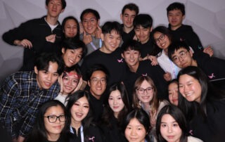 A group of young people, participants of a student-run hackathon, pose closely together, smiling at the camera. Clad in casual attire with some sporting matching black tops featuring a small pink logo, they stand against a simple, light-colored wall celebrating their Geese Hacks experience.