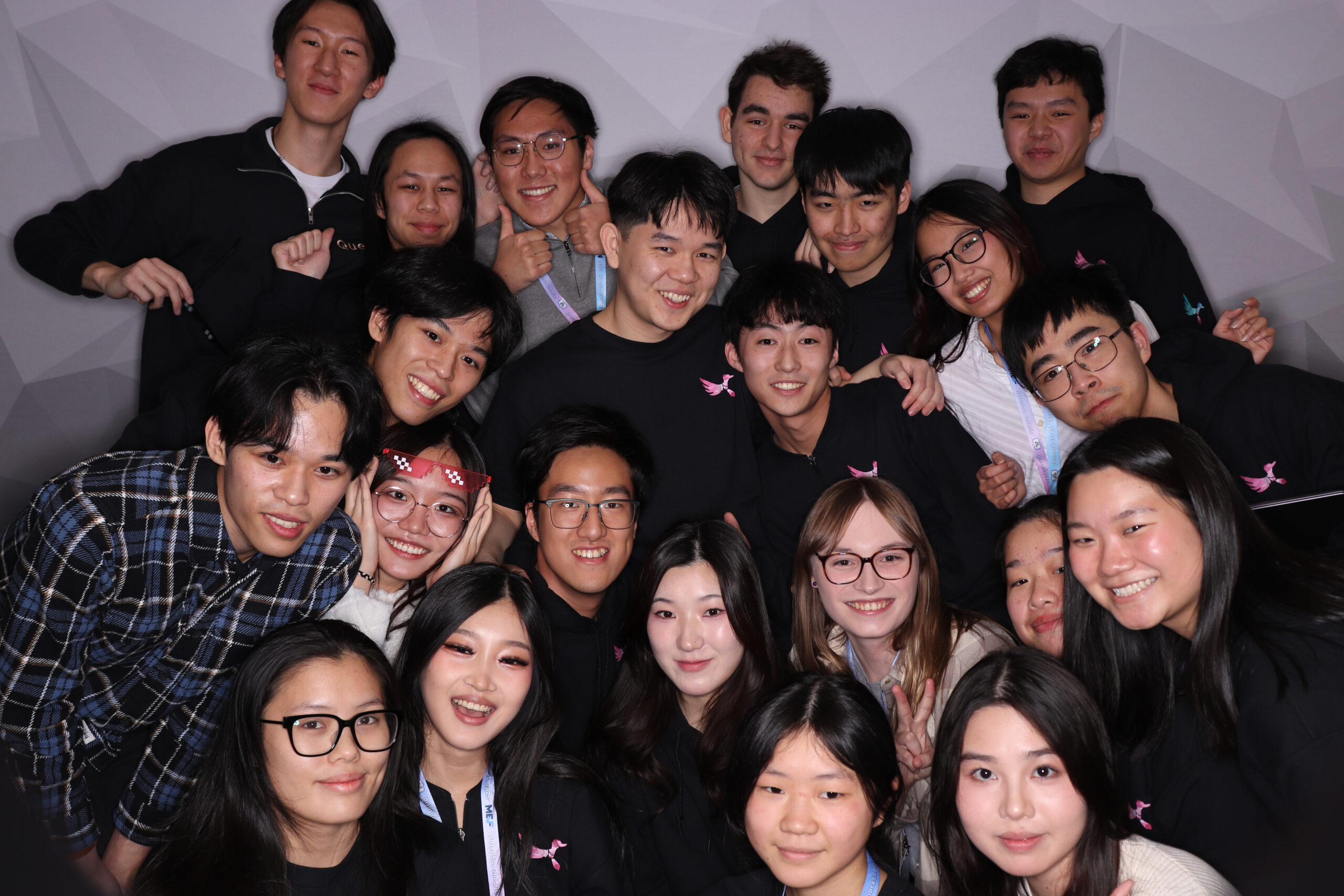 A group of young people, participants of a student-run hackathon, pose closely together, smiling at the camera. Clad in casual attire with some sporting matching black tops featuring a small pink logo, they stand against a simple, light-colored wall celebrating their Geese Hacks experience.