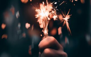 A hand holds a lit sparkler, emitting bright, fiery sparks against a dark background. The sparks create a dynamic and festive atmosphere, reminiscent of New Year's Day celebrations, with small particles glowing and scattering around the sparkler.