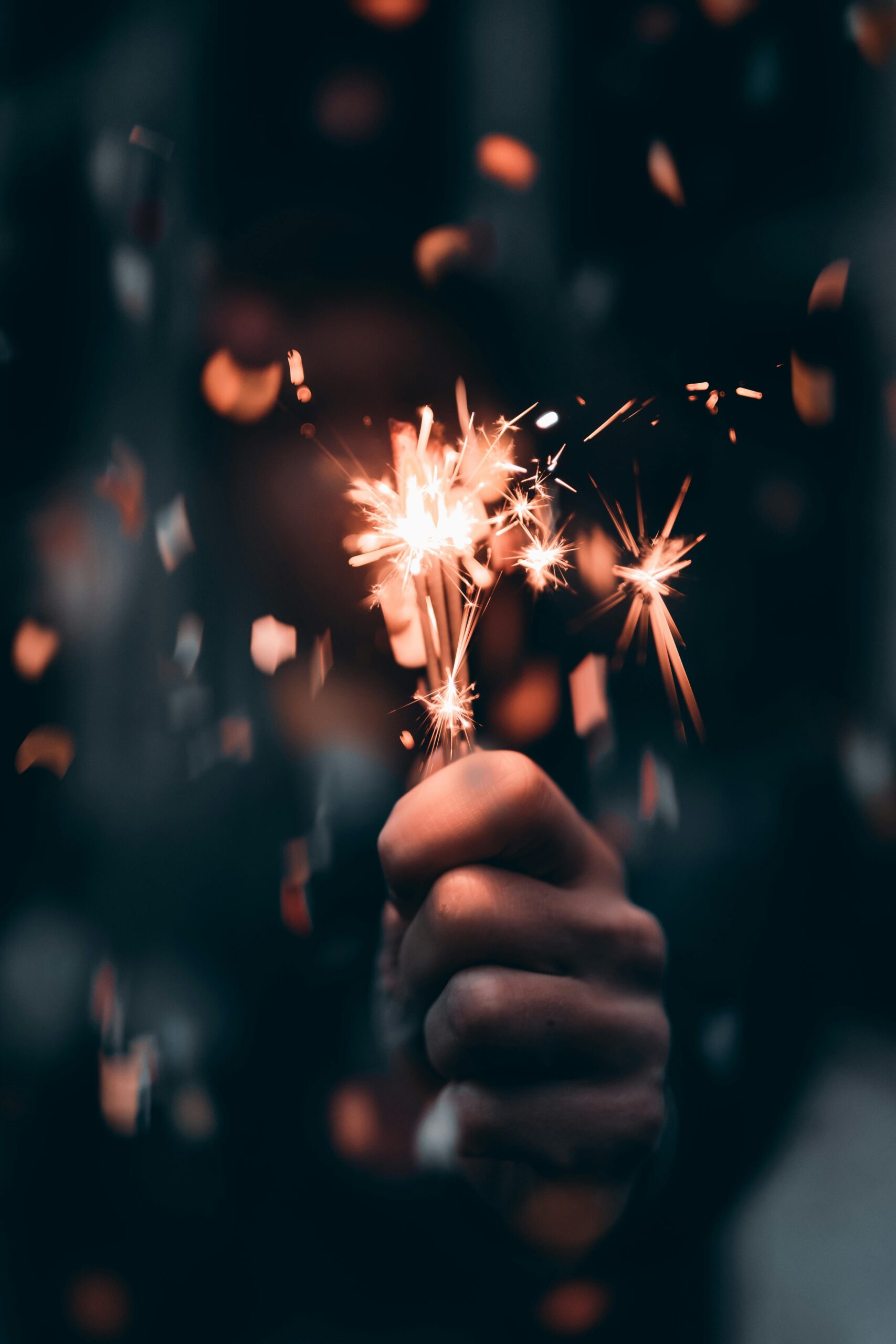 A hand holds a lit sparkler, emitting bright, fiery sparks against a dark background. The sparks create a dynamic and festive atmosphere, reminiscent of New Year's Day celebrations, with small particles glowing and scattering around the sparkler.