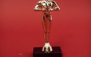 A shiny gold statuette resembling an Academy Award stands on a black base against a red background, with small gold stars scattered around, evoking anticipation for early Oscars 2025 predictions.