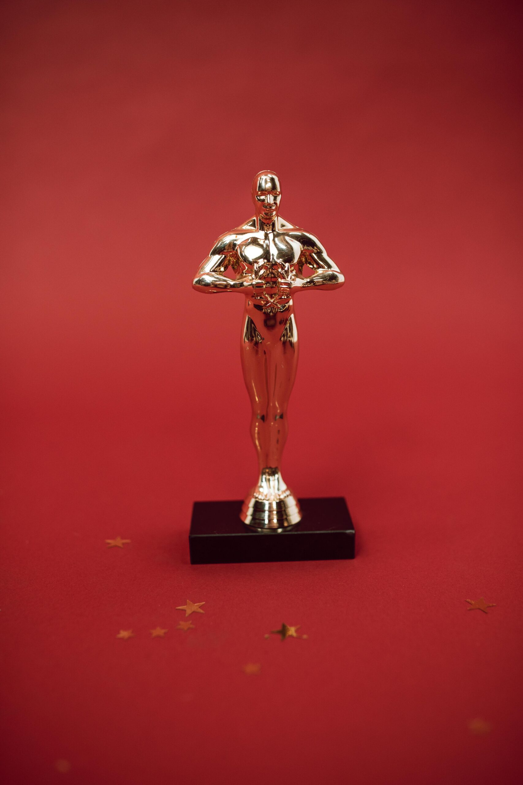 A shiny gold statuette resembling an Academy Award stands on a black base against a red background, with small gold stars scattered around, evoking anticipation for early Oscars 2025 predictions.