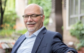 A smiling man with glasses, an engineering alumnus from Waterloo, sits outdoors on a bench in a suit jacket over a checkered shirt. Blurred greenery and building facades frame the peaceful, natural setting.