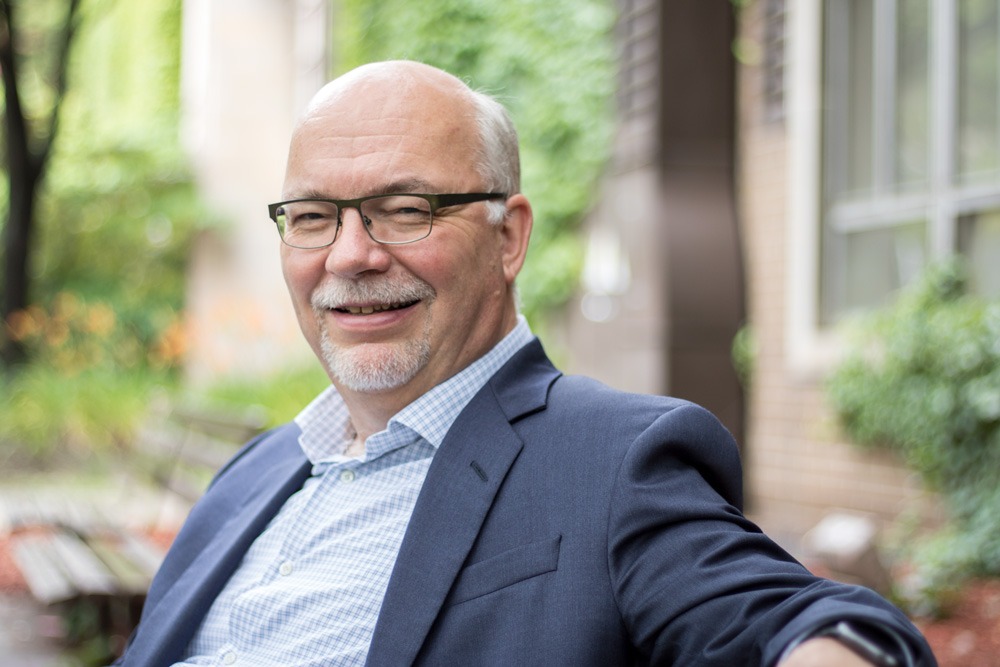 A smiling man with glasses, an engineering alumnus from Waterloo, sits outdoors on a bench in a suit jacket over a checkered shirt. Blurred greenery and building facades frame the peaceful, natural setting.