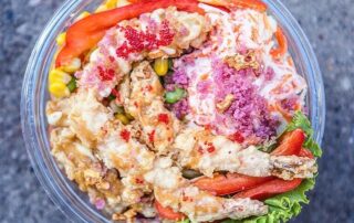 Join the Poke Party with a vibrant poke bowl from Rolltation, featuring tempura shrimp, bright red fish roe, shredded vegetables, corn, sliced red bell peppers, and crispy toppings. This colorful meal-deal is beautifully arranged on a bed of lettuce in a clear bowl.