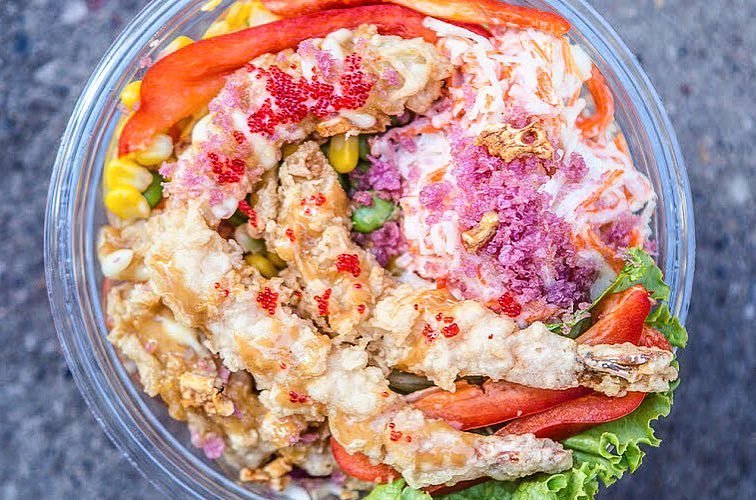 Join the Poke Party with a vibrant poke bowl from Rolltation, featuring tempura shrimp, bright red fish roe, shredded vegetables, corn, sliced red bell peppers, and crispy toppings. This colorful meal-deal is beautifully arranged on a bed of lettuce in a clear bowl.