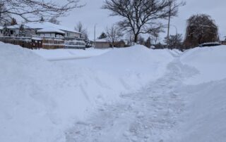 A snow-covered residential street in Waterloo features high snowbanks from a recent snowstorm on either side. The path is shoveled, providing clear walking space. Bare trees and houses with snow-covered roofs line the street under a cloudy sky, hinting at the extreme cold.