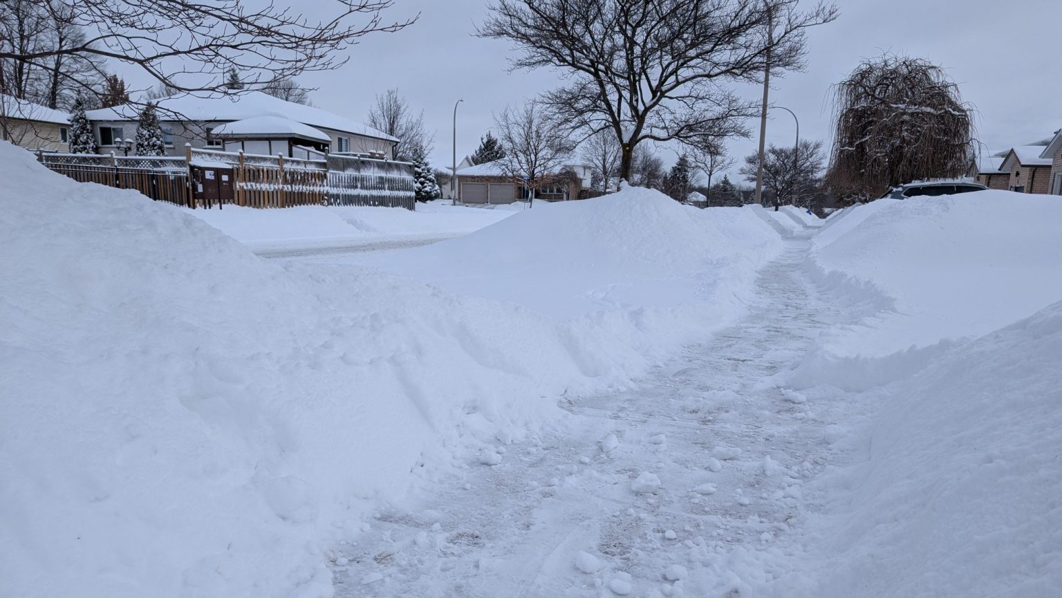 A snow-covered residential street in Waterloo features high snowbanks from a recent snowstorm on either side. The path is shoveled, providing clear walking space. Bare trees and houses with snow-covered roofs line the street under a cloudy sky, hinting at the extreme cold.