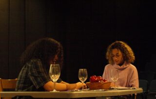 Two people with curly hair sit across from each other at a table with empty wine glasses and a bowl of apples, reminiscent of a scene from an Upstart Festival performance. They appear focused, set against a dimly lit background.