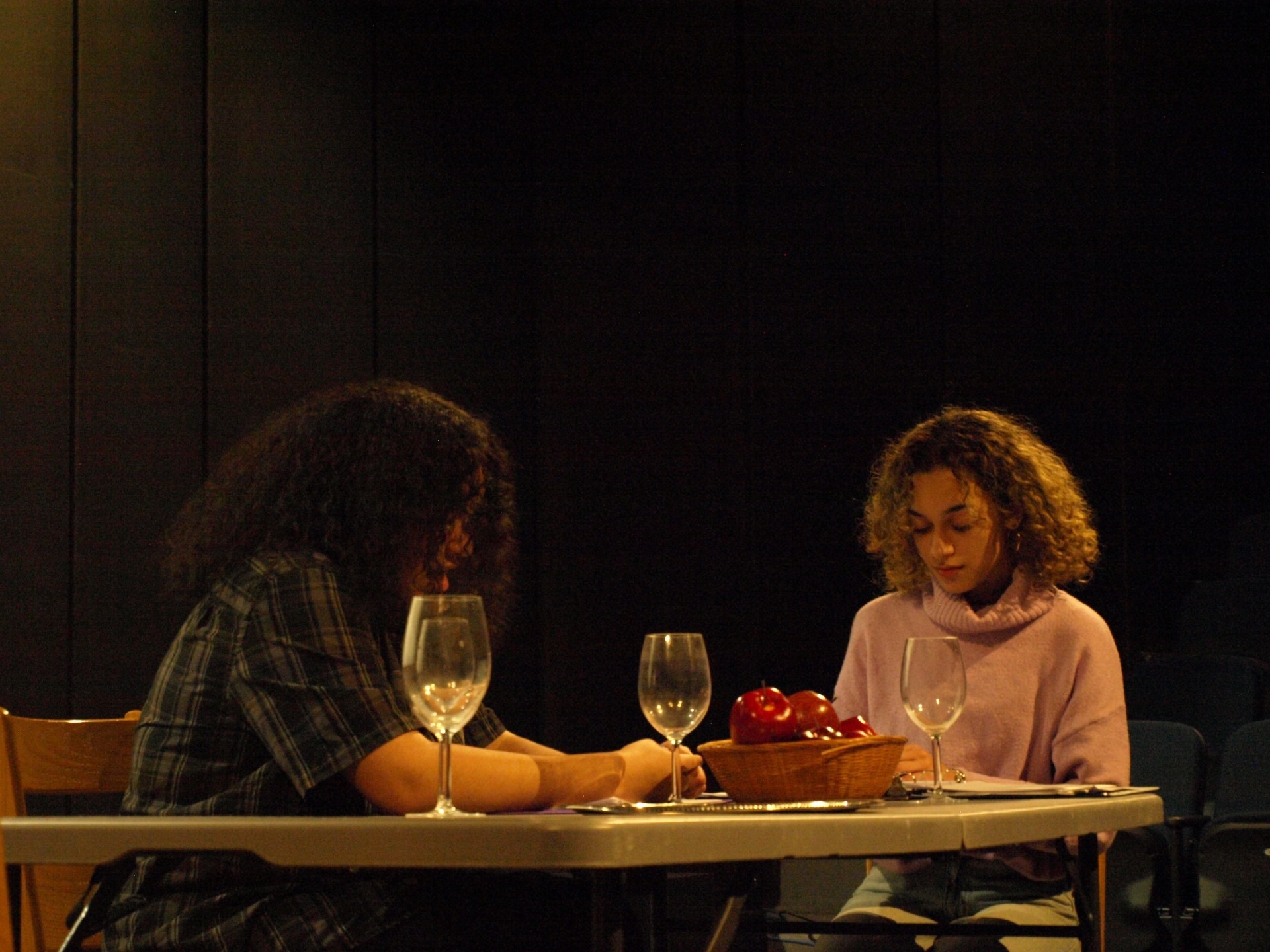 Two people with curly hair sit across from each other at a table with empty wine glasses and a bowl of apples, reminiscent of a scene from an Upstart Festival performance. They appear focused, set against a dimly lit background.