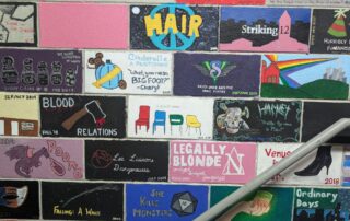 A colorful wall mural celebrates 90 years of KWLT, spotlighting various theater production titles and themes in rectangular blocks. The artwork includes text like "Hair," "Legally Blonde," and "Cinderella," alongside bright visuals and abstract designs. A metal handrail runs across.