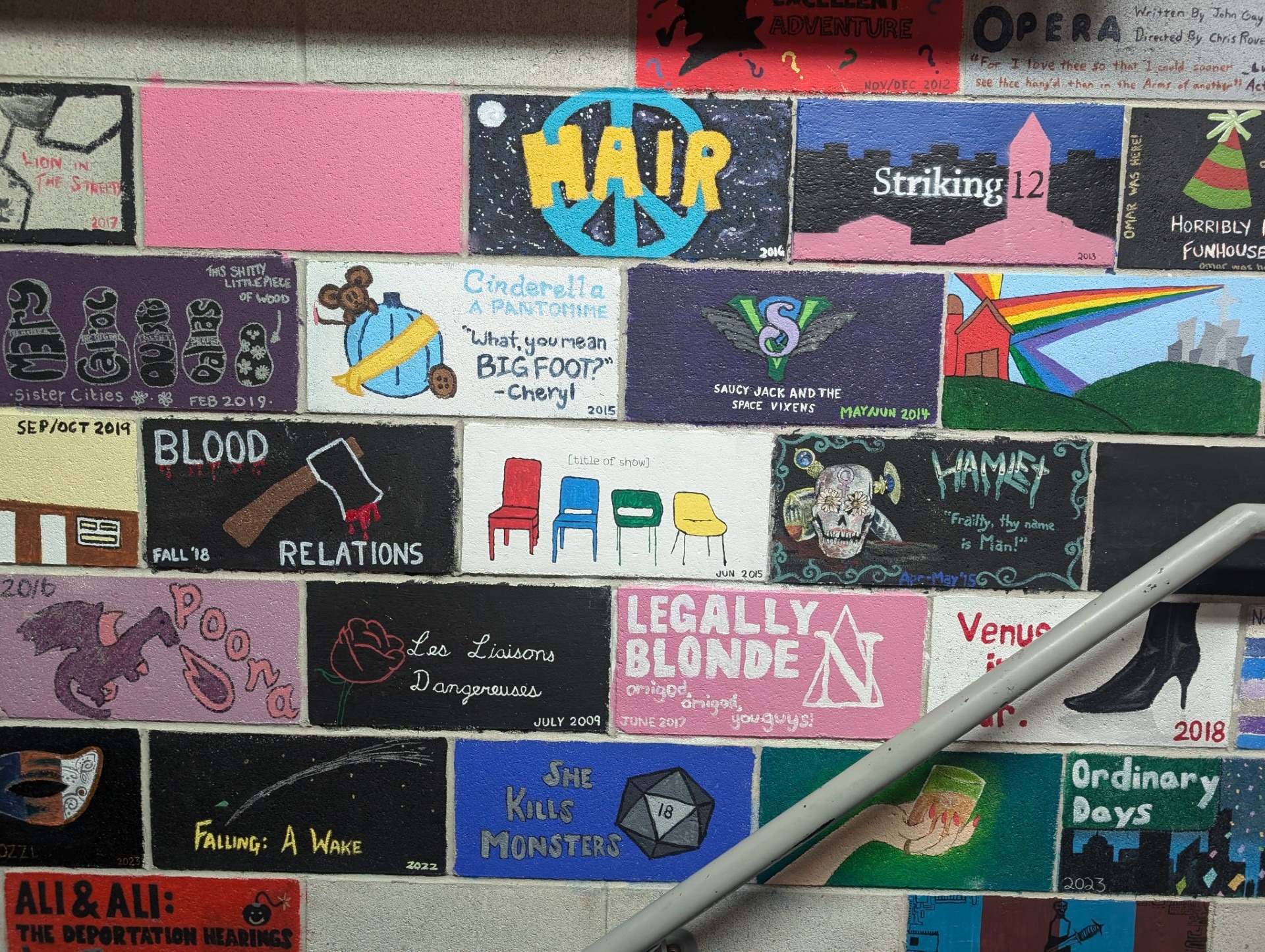 A colorful wall mural celebrates 90 years of KWLT, spotlighting various theater production titles and themes in rectangular blocks. The artwork includes text like "Hair," "Legally Blonde," and "Cinderella," alongside bright visuals and abstract designs. A metal handrail runs across.