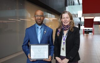 A man in a blue suit holds a framed Equity and Inclusivity Award, standing beside a woman in a black blazer and colorful scarf. They are in a modern building with glass walls and red accents. Both are smiling at the camera.