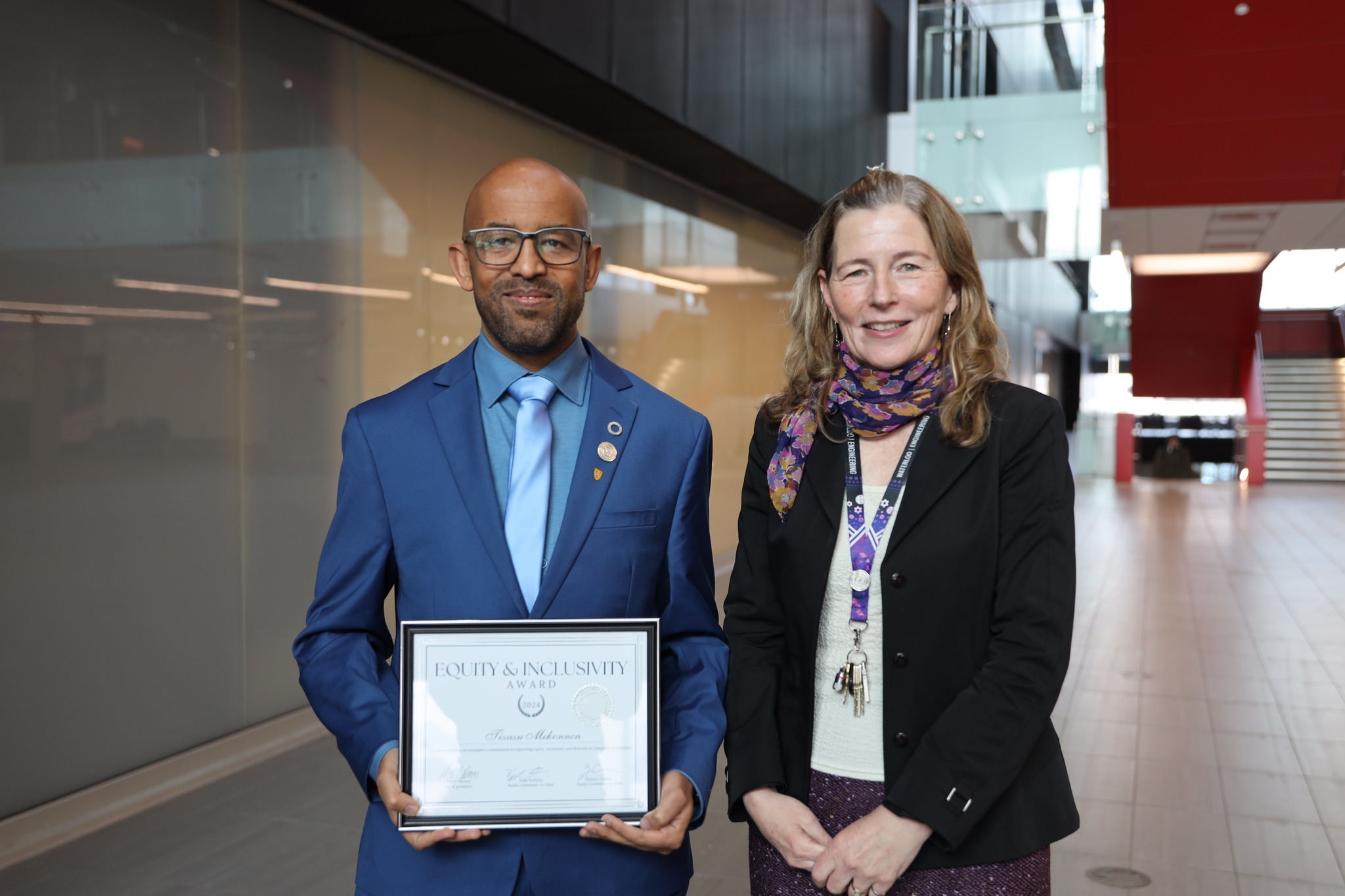 A man in a blue suit holds a framed Equity and Inclusivity Award, standing beside a woman in a black blazer and colorful scarf. They are in a modern building with glass walls and red accents. Both are smiling at the camera.