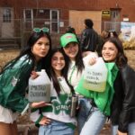 Four women celebrating St. Patrick's Day, wearing festive green attire. Two are holding jugs labeled "Metro Borgin" and "It's Borg o'clock somewhere." A crowd in similar attire is in the background near brick buildings.