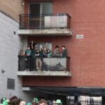 A group of people wearing green celebrate on a balcony of a brick building. Some wave at the camera, while others smile. Below them, a crowd of people wearing green hats is partially visible.