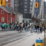 A group of people, many wearing green, walk across a street in an urban area. Buildings, including a red house and taller structures, are in the background. Traffic lights are visible, and it's a cloudy day.