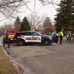 A police SUV and a fire truck block a road lined with people wearing green shirts. Officers stand nearby directing the crowd. Trees and houses are in the background, and it's a cloudy day.