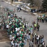 A large crowd of people dressed in green gather on a street, likely for a celebration or parade. Some police officers stand nearby as others observe the event. Trees and patches of snow can be seen, suggesting it's early spring.