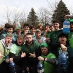 A group of young people wearing green outfits and accessories, some holding large bottles of liquid, celebrate outdoors. They pose enthusiastically, with several making playful gestures. Trees and a cloudy sky are in the background.