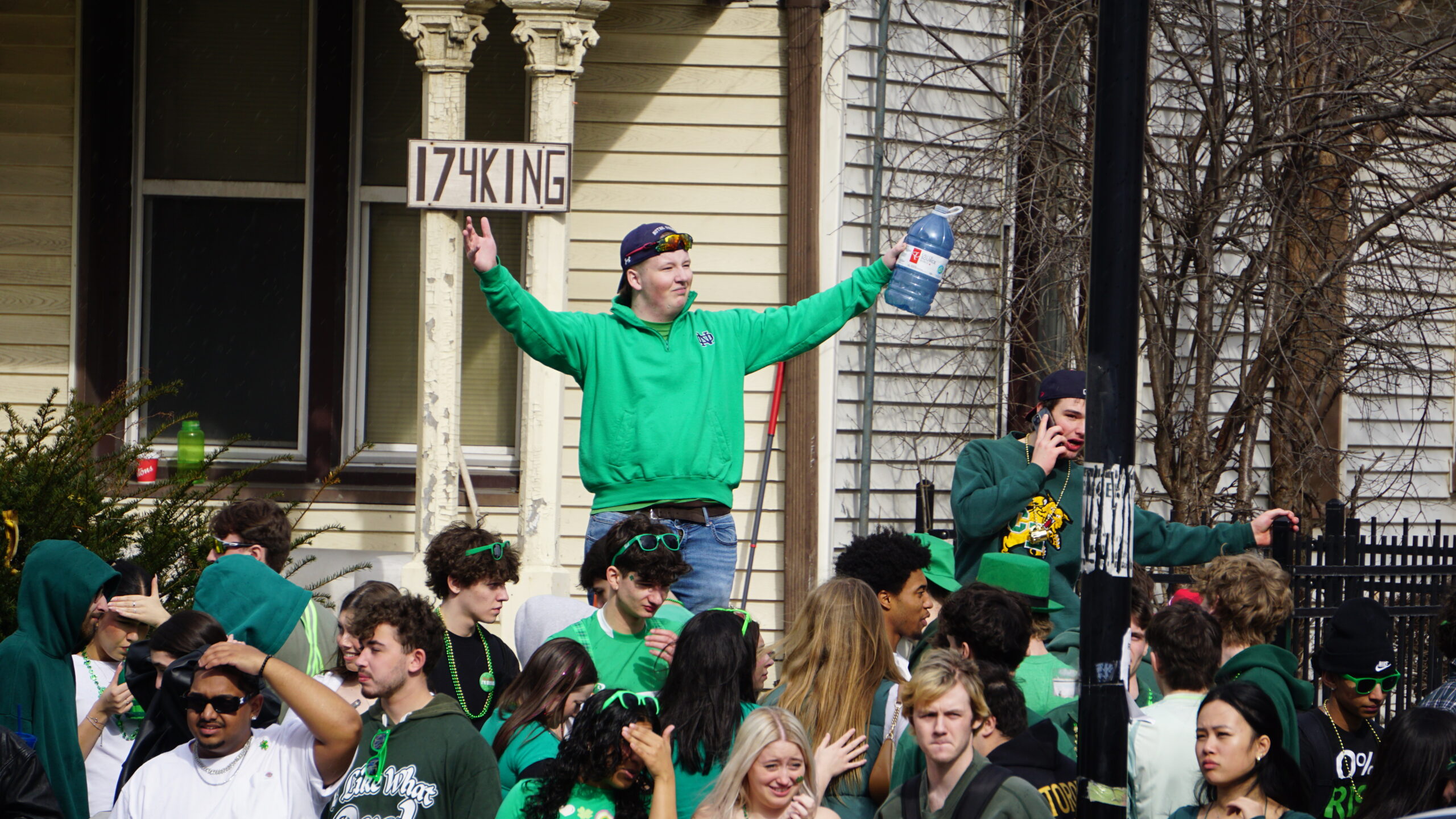 A group of people celebrate St. Patrick's Day outdoors, mostly dressed in green. One person stands elevated with outstretched arms holding a bottle. The scene is festive, with decorations and a street sign in the background reading "17 KING," reminiscent of Waterloo's vibrant spirit.