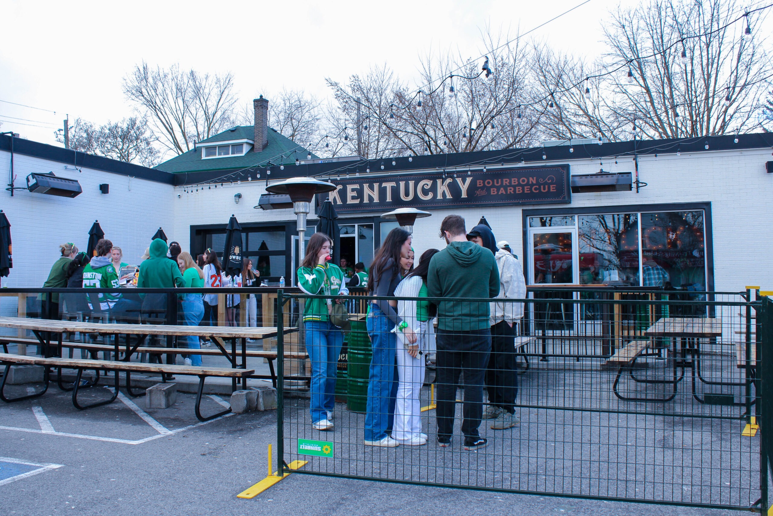 A group of people in green clothing gathers outside "Kentucky Bourbon & Barbecue" in uptown Waterloo on a cloudy day. They're standing by picnic tables and outdoor heaters under string lights, with bare trees and a house in the background.